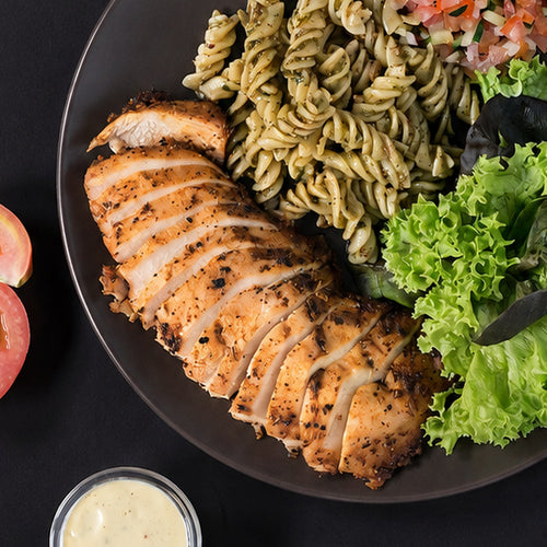 Sliced Cajun-roasted chicken breast, photographed on a dark plate with pesto fusilli and lettuce. Serving suggestion.